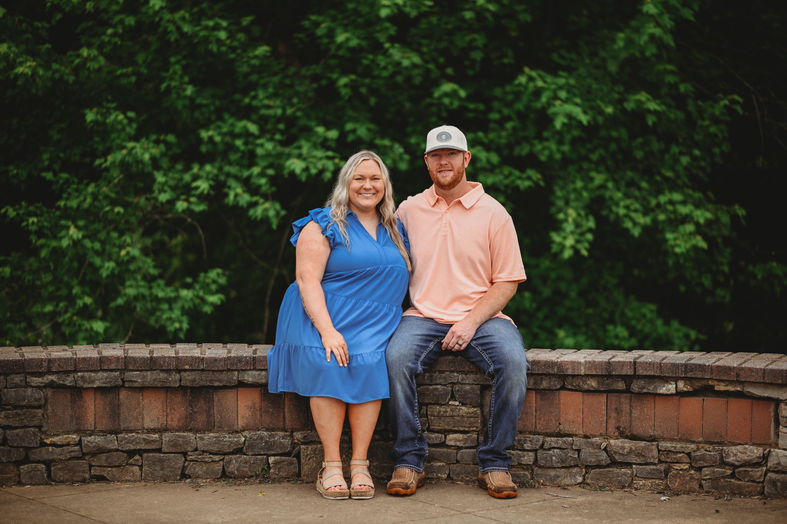 A couple sitting together on a stone wall with trees in the background.