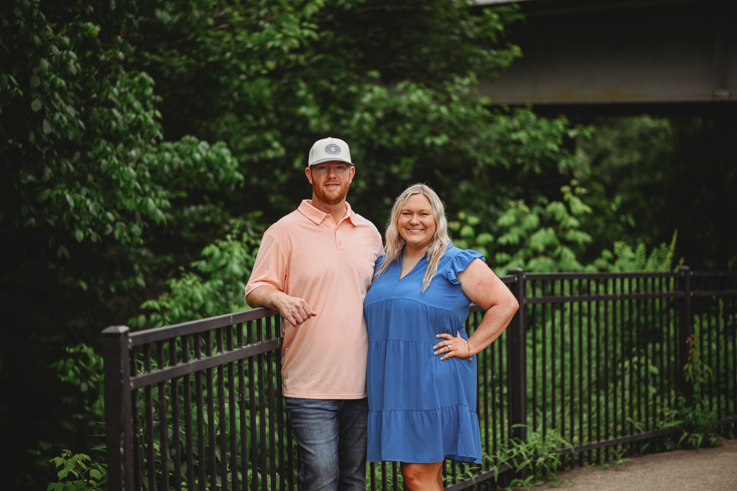 A couple posing against a metal railing with green foliage in the background.