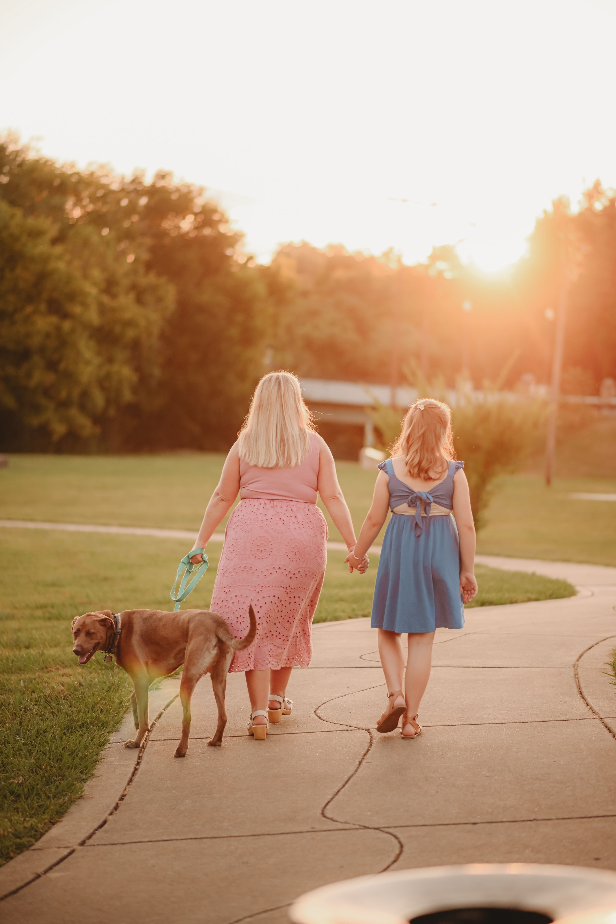 Two people and a dog walking on a paved path at sunset.