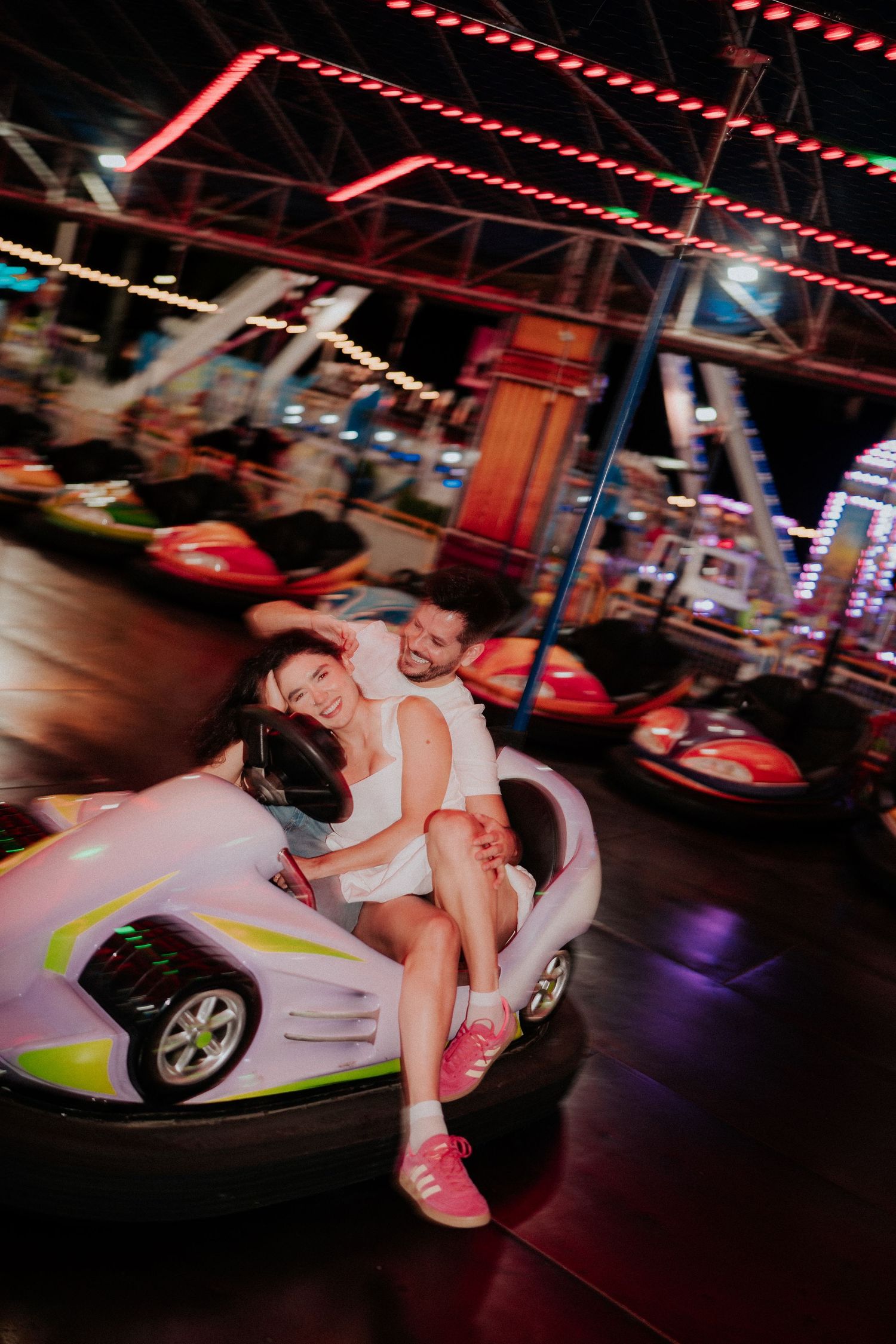 Couple enjoys a romantic moment on bumper cars at an amusement park with red neon lights overhead.