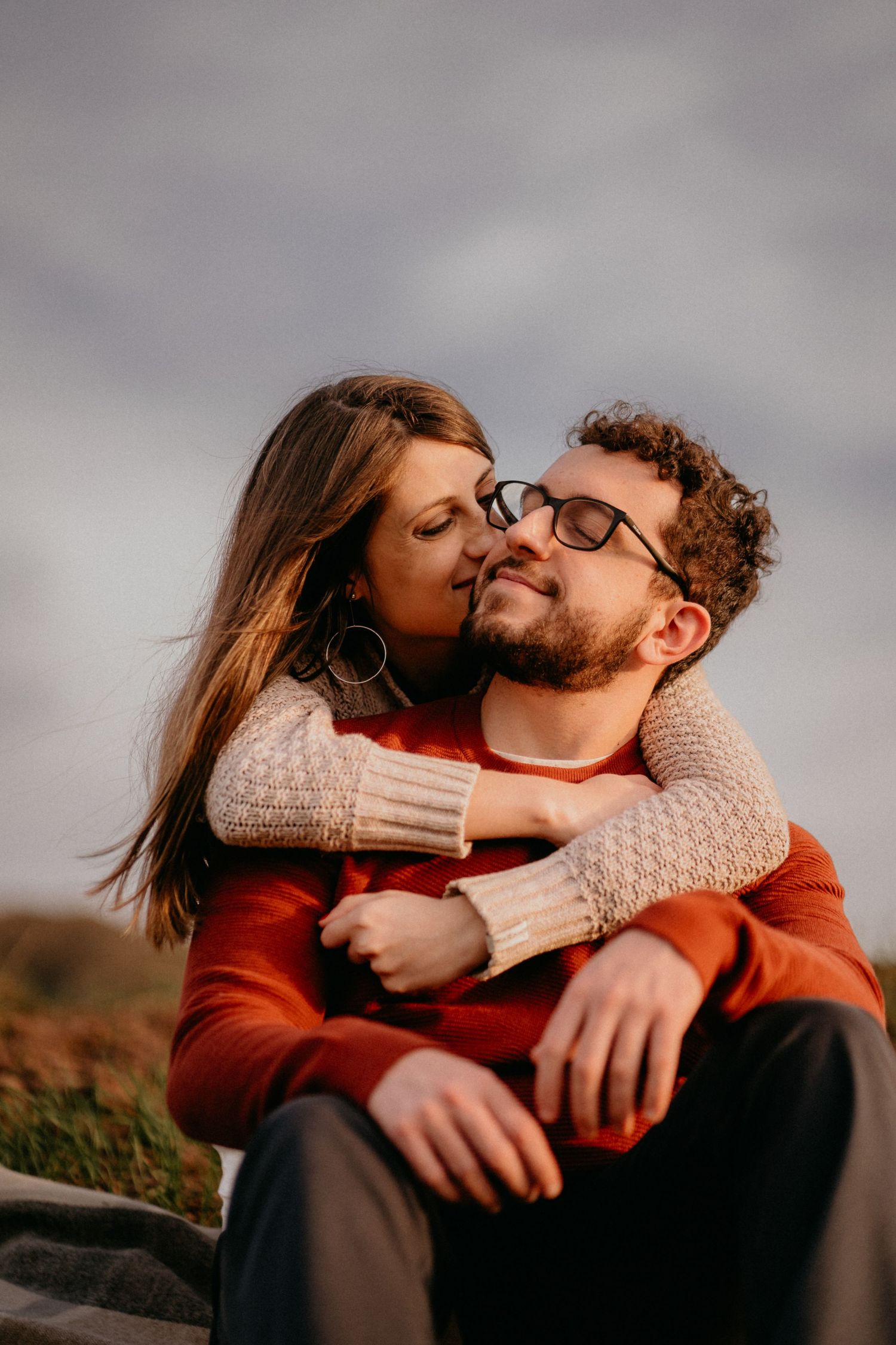 Intimate moment between couple wearing cozy sweaters during sunset photoshoot.