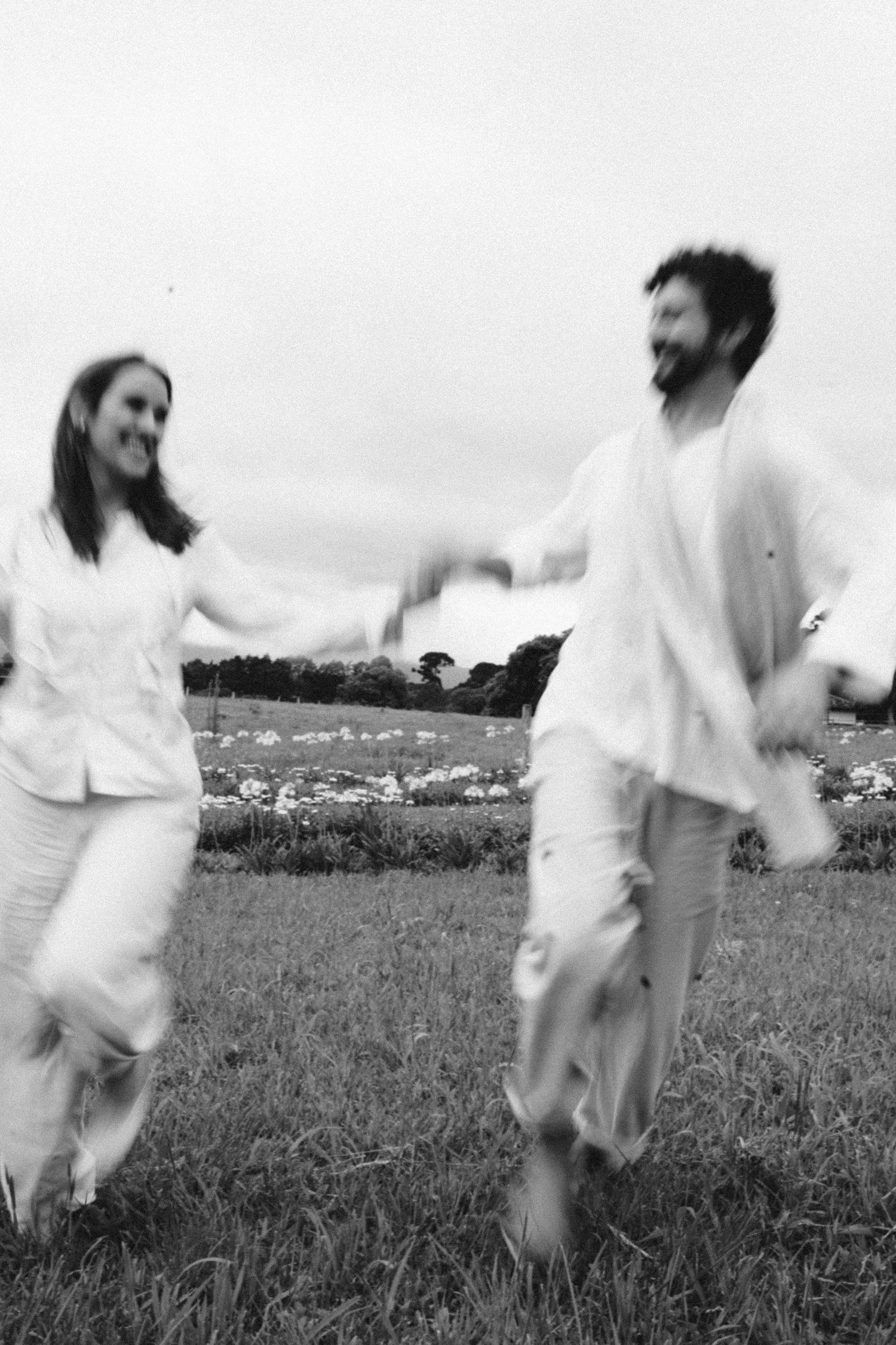 Black and white photo of couple running joyfully through field in white clothing.