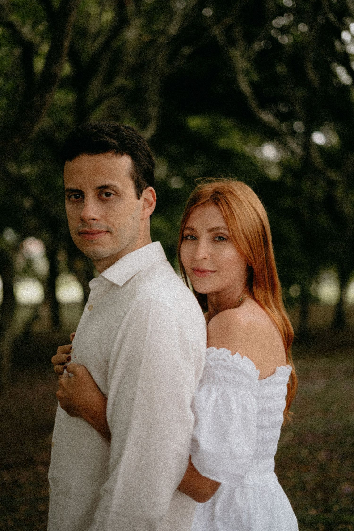 Romantic couple in white outfits embrace during outdoor photoshoot with trees in background.