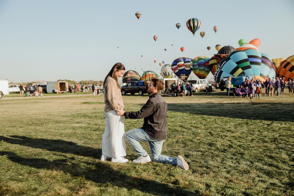 Man proposing on one knee to woman at a hot air balloon festival on a sunny day.