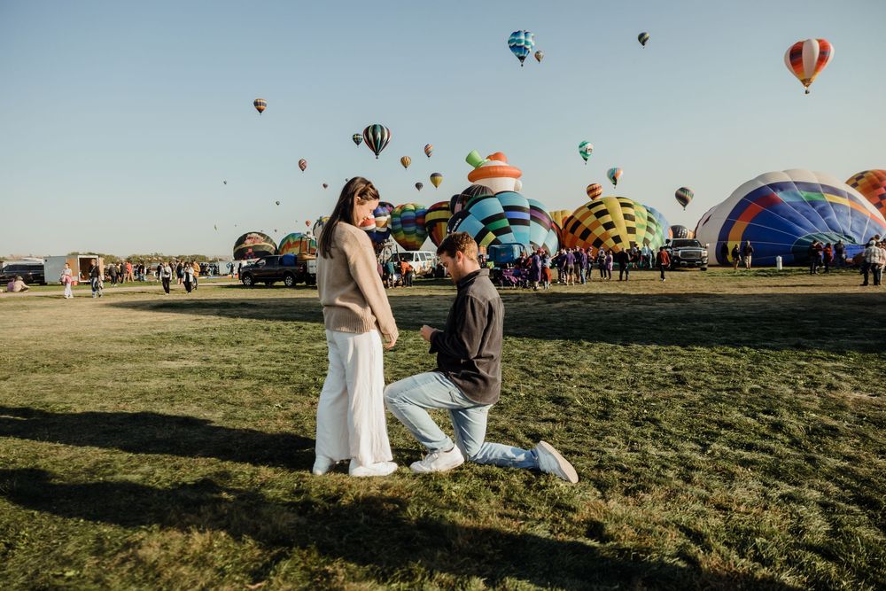 Man kneeling in proposal to woman surrounded by colorful hot air balloons at a festival.