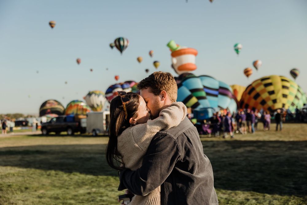 Couple sharing a kiss at a hot air balloon festival with colorful balloons in background.