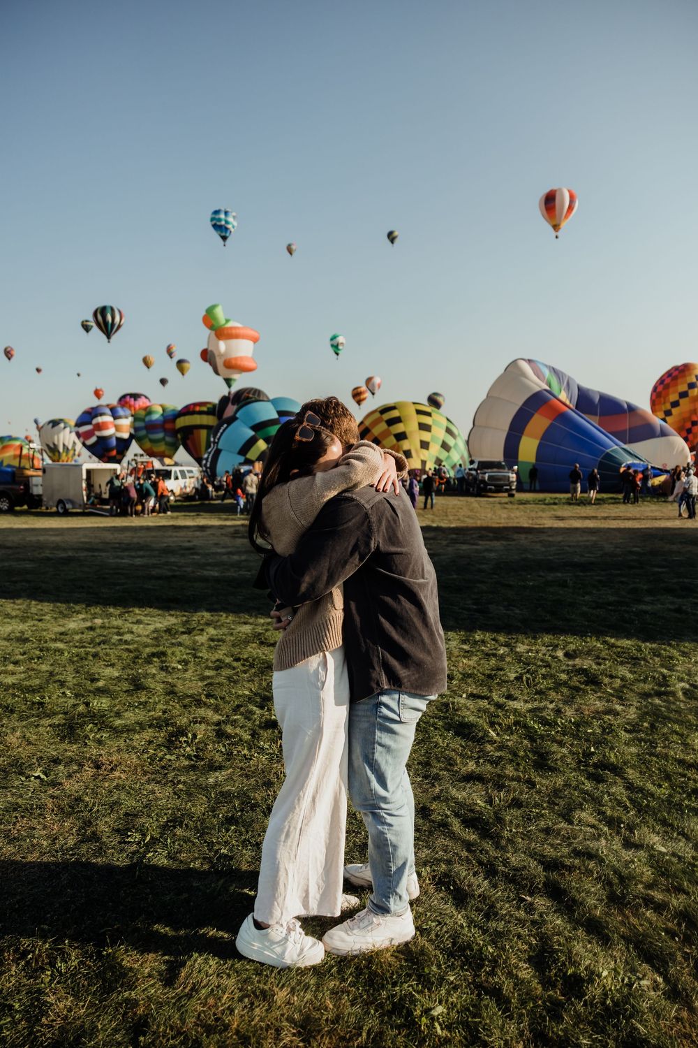 Engaged couple hugging while surrounded by colorful hot air balloons launching into the sky.