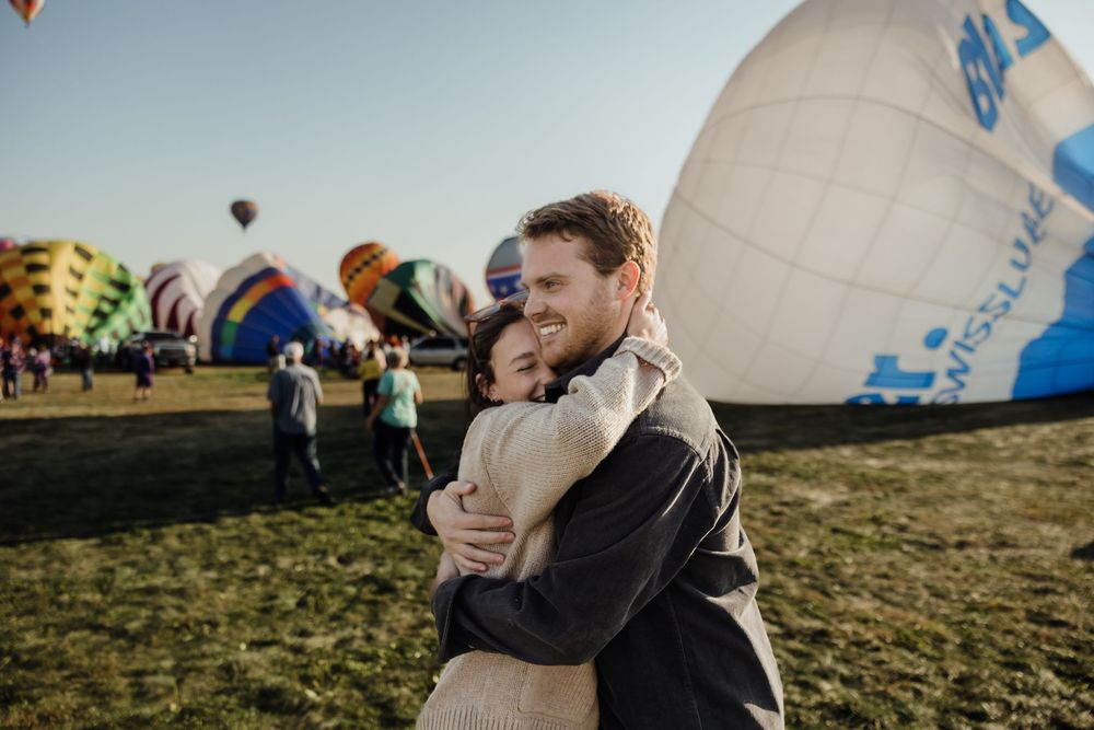 Happy couple embracing and smiling at a hot air balloon festival on a sunny morning.