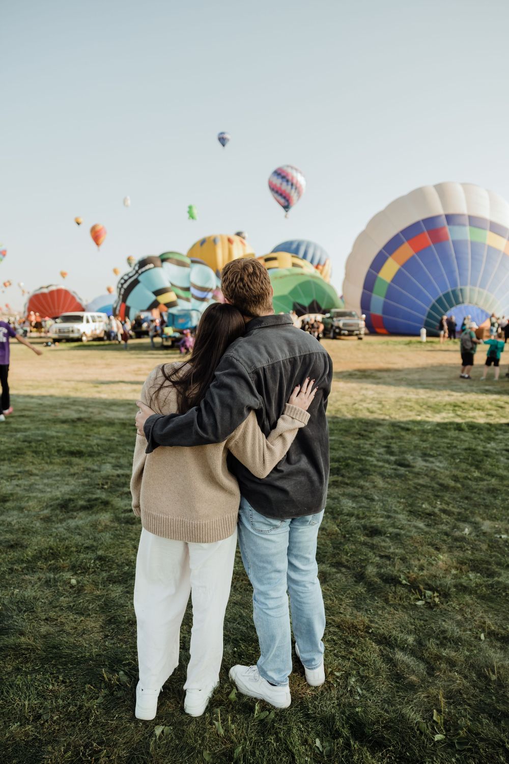 Couple embracing from behind while watching colorful hot air balloons launch at a festival.