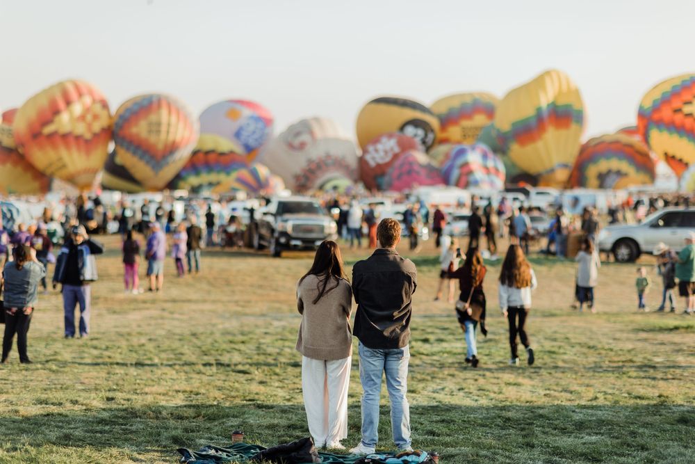 Couple stands arm in arm on a grassy field watching dozens of colorful hot air balloons at a festival.