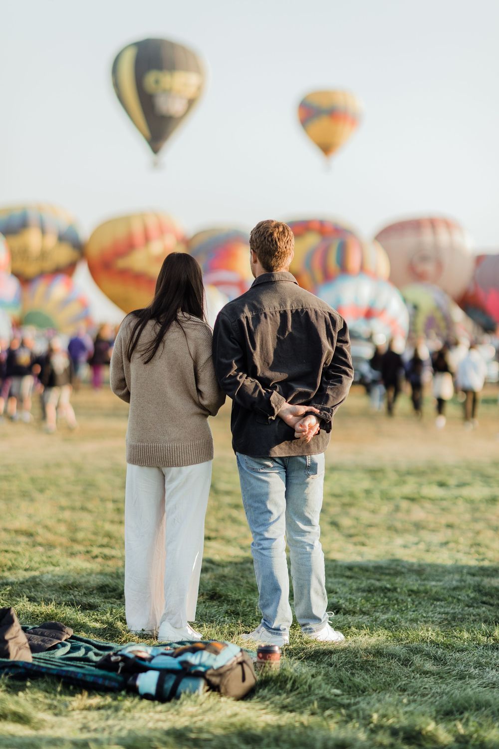 Couple holds hands from behind while watching colorful hot air balloons fill the sky at an outdoor festival.