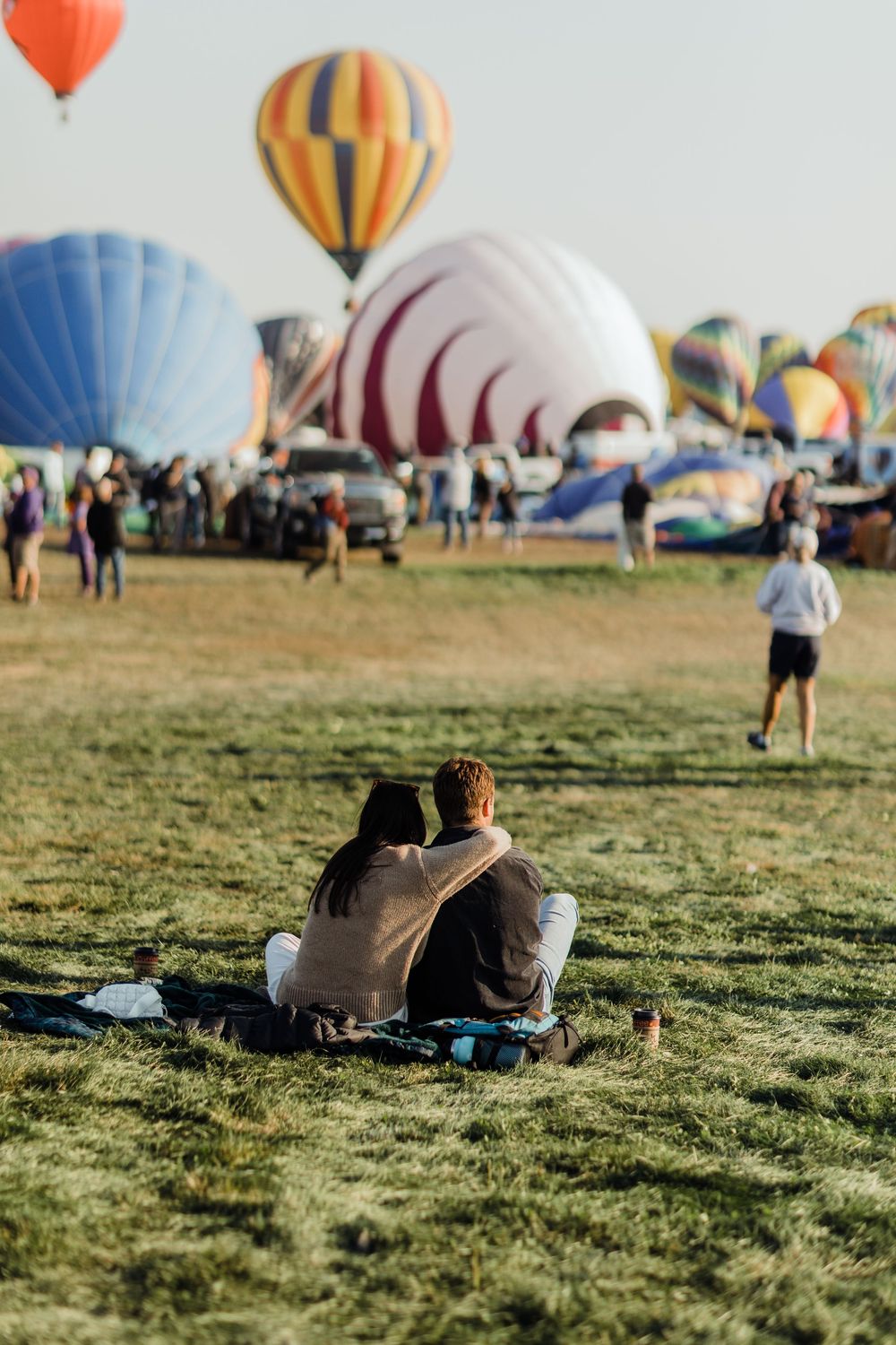 Couple sitting on grass watching colorful hot air balloons inflate at an outdoor festival.