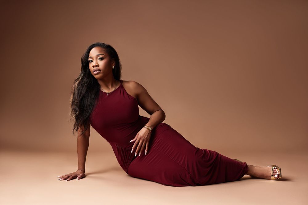 Woman in burgundy dress poses elegantly against warm-toned backdrop in glamour photoshoot series.