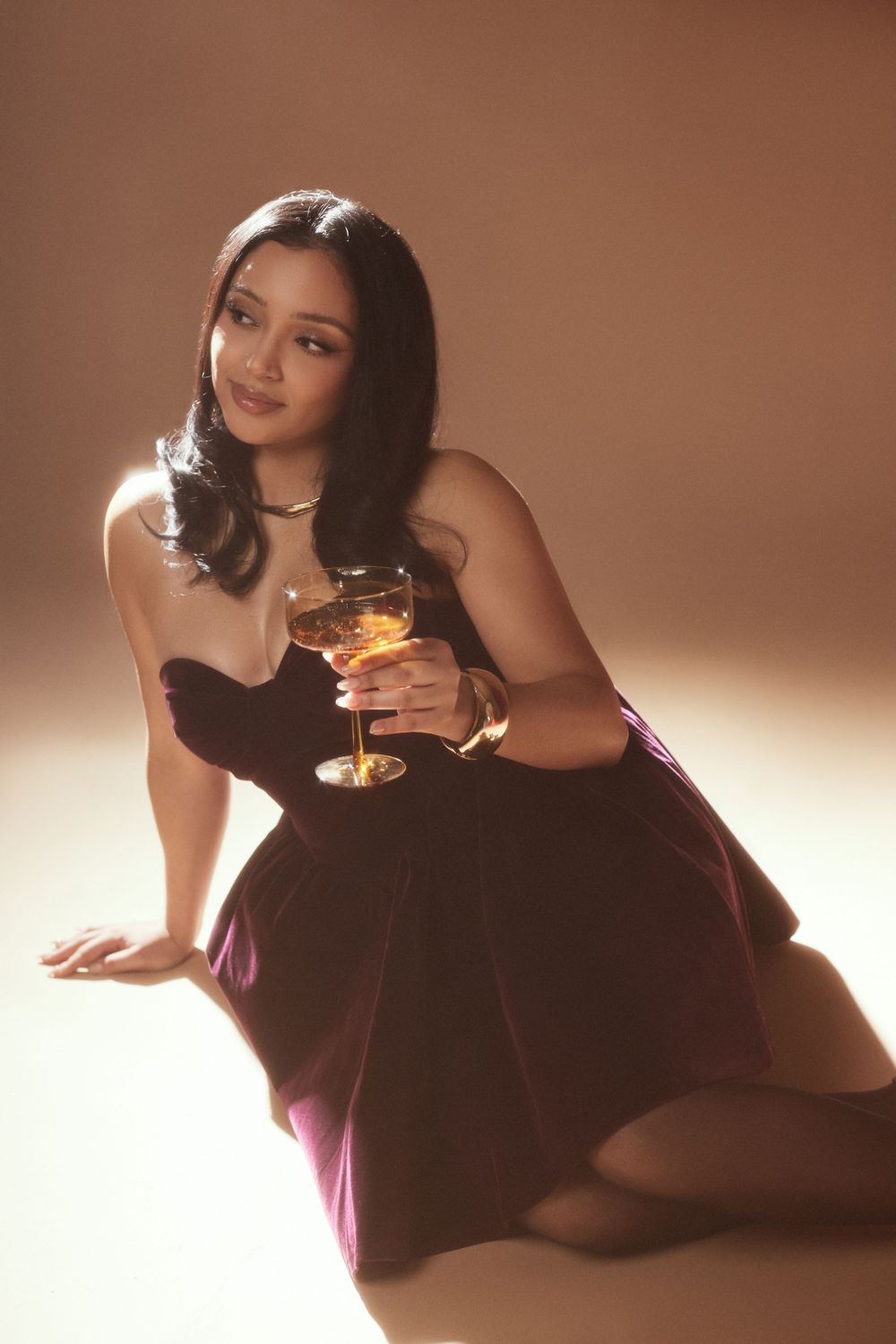 Elegant photoshoot featuring burgundy dress and cocktail glass in warm lighting.