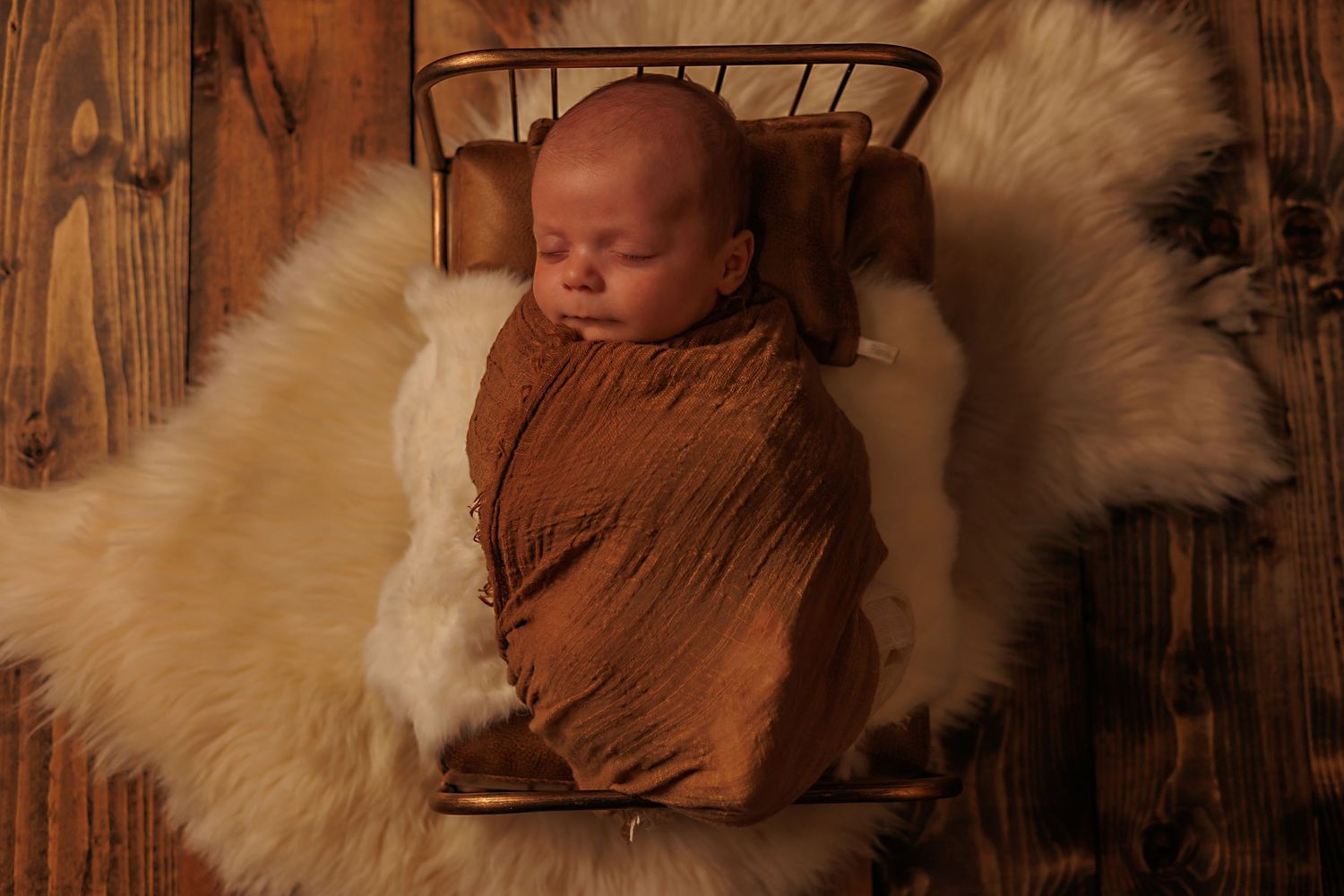 A sleeping baby wrapped in brown fabric rests on white fur against a wooden backdrop.
