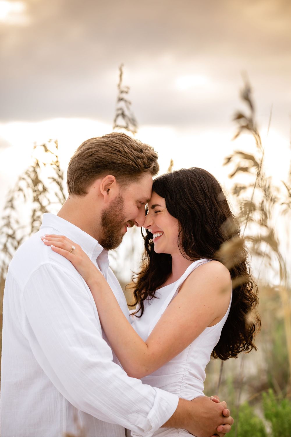 A romantic couple in white clothing shares tender moments at sunset in a field of tall grass and wildflowers.