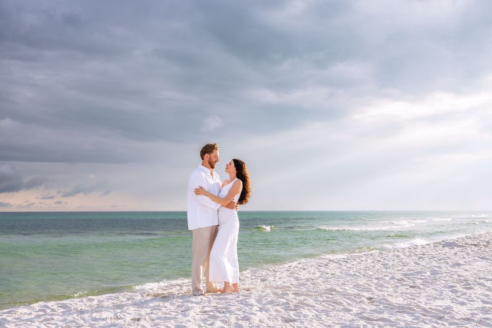 Romantic couple embracing on a serene white sandy beach with turquoise waters and dramatic cloudy skies.