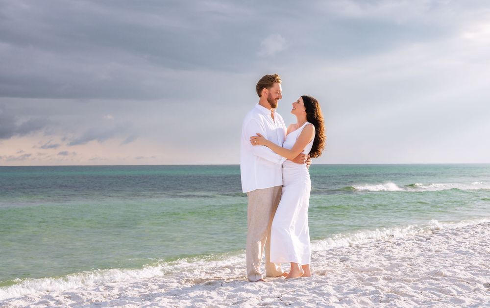 Two people in white outfits share a romantic moment on a beach with waves crashing in the background.