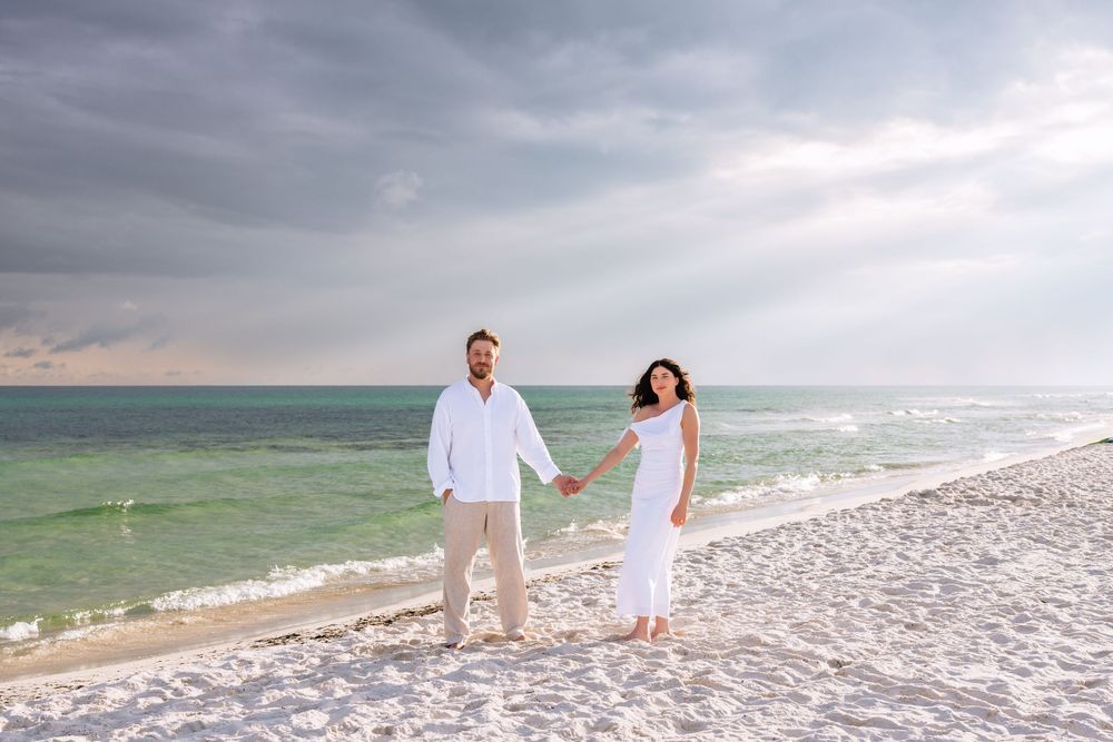 People walking along a scenic beach shoreline at sunset with waves rolling onto white sand under cloudy skies.