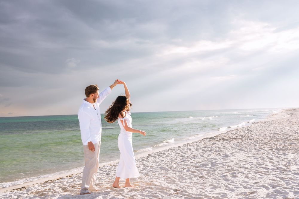 Two figures in white outfits walk together peacefully along a pristine beach with turquoise waters and cloudy skies.