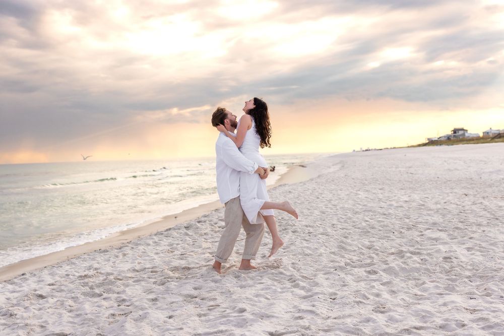 Two people share a romantic moment on a white sandy beach at sunset with ocean waves in the background.