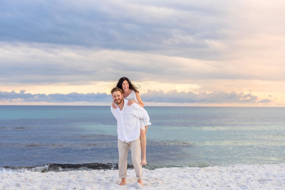 Couple in white attire shares a romantic moment on a beach at sunset with ocean and cloudy sky in background.