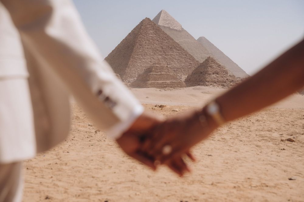 Hands holding and releasing sand in a desert landscape with pyramids visible in the background.