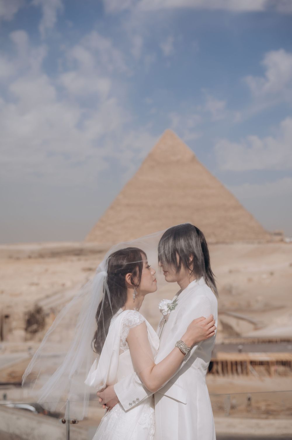 Two people in white outfits embrace with ancient Egyptian pyramids in the background under a cloudy sky.