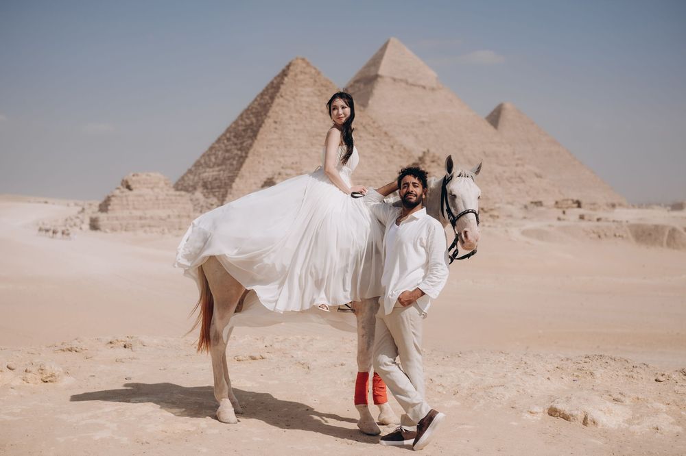 A couple in wedding attire pose with a white horse in front of the ancient Egyptian pyramids of Giza in the desert.