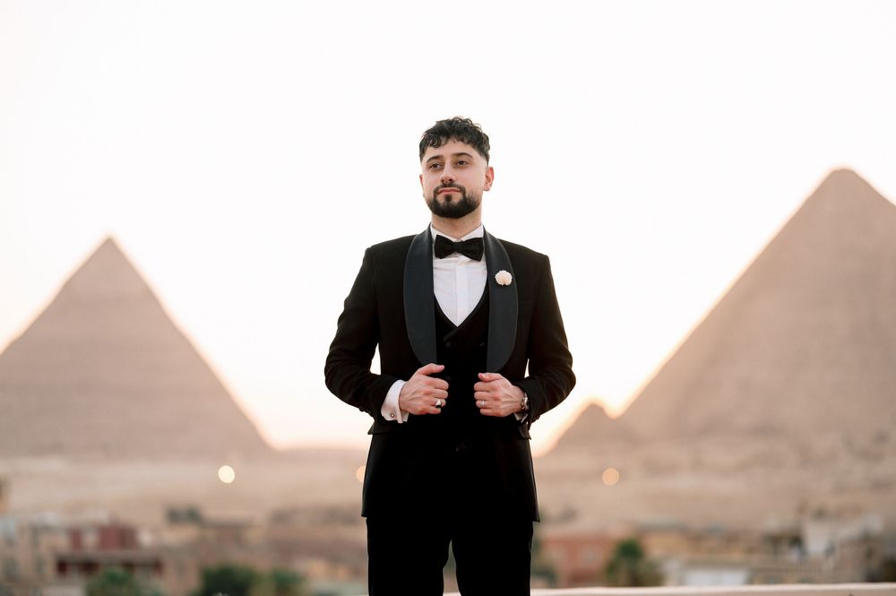 A groom in a black tuxedo and bow tie poses in front of the ancient pyramids of Giza at sunset.