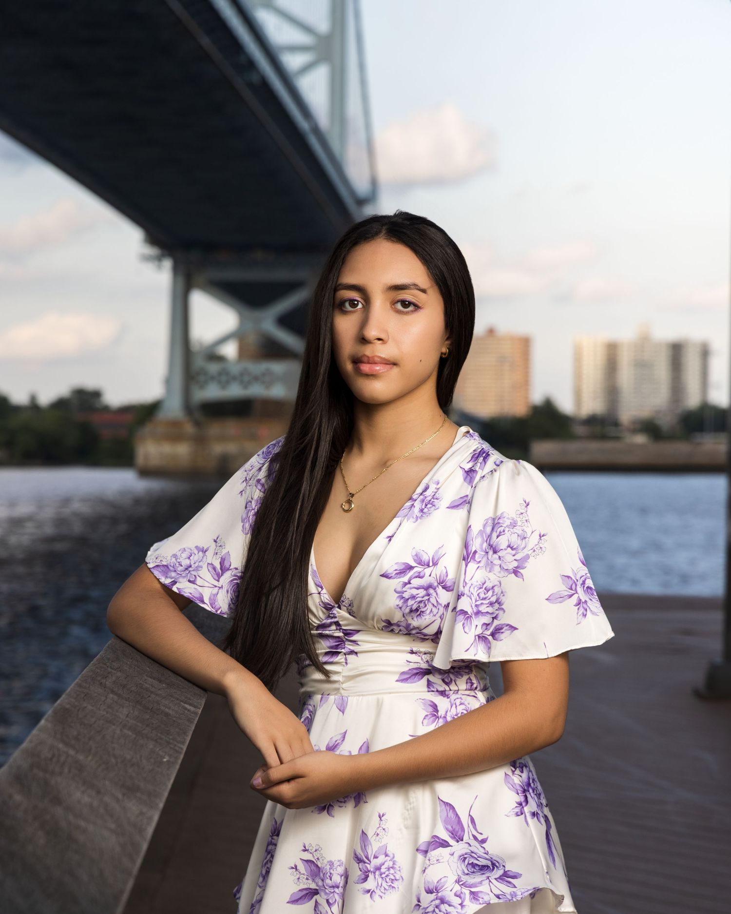 A woman in a floral white and purple dress sits on a wooden boardwalk during sunset near a harbor.