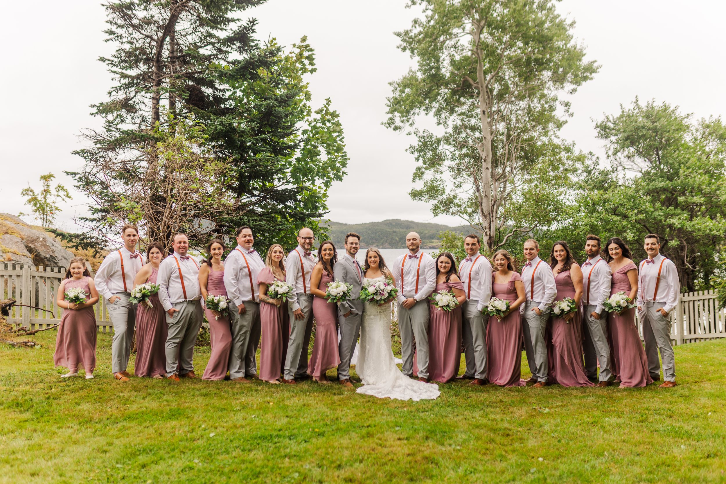 Wedding party in burgundy dresses and suits pose together in a field with trees in the background during golden hour.