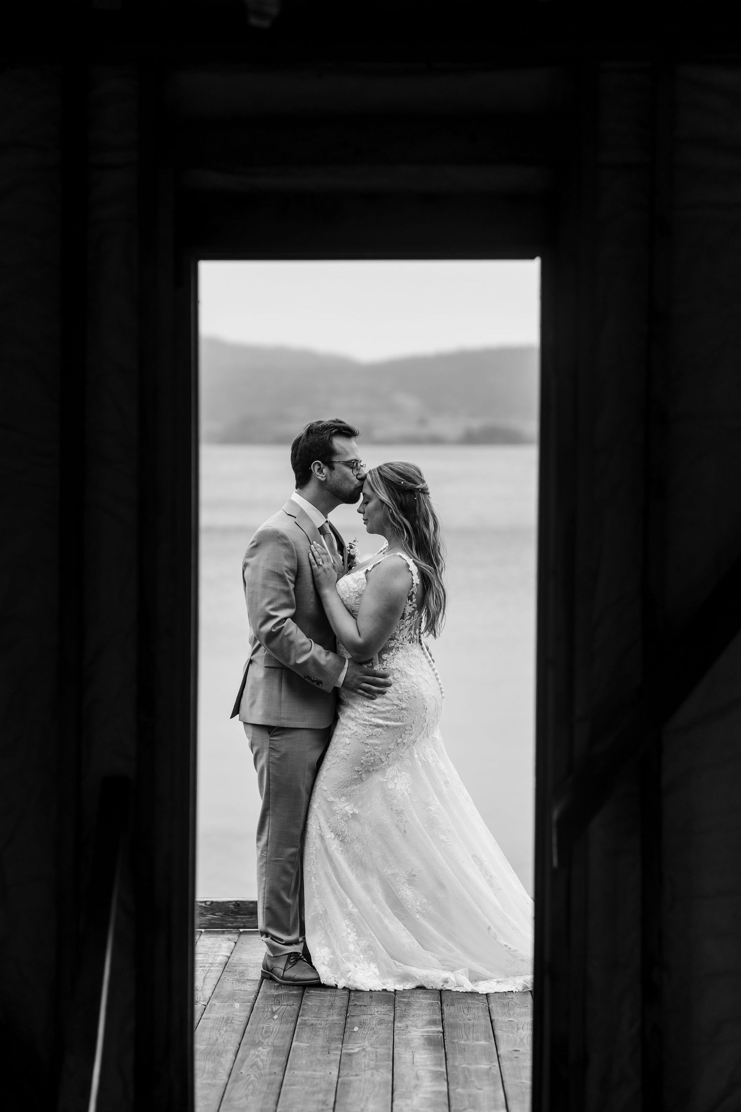 A romantic black and white photo series shows a couple kissing on a dock by a lake in their wedding attire.