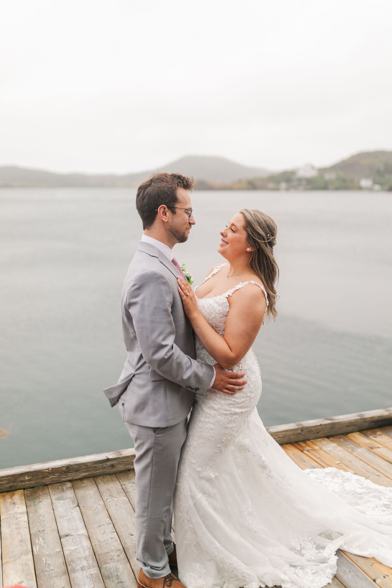 A couple shares a romantic dance moment together on a waterfront dock overlooking misty mountains and water.