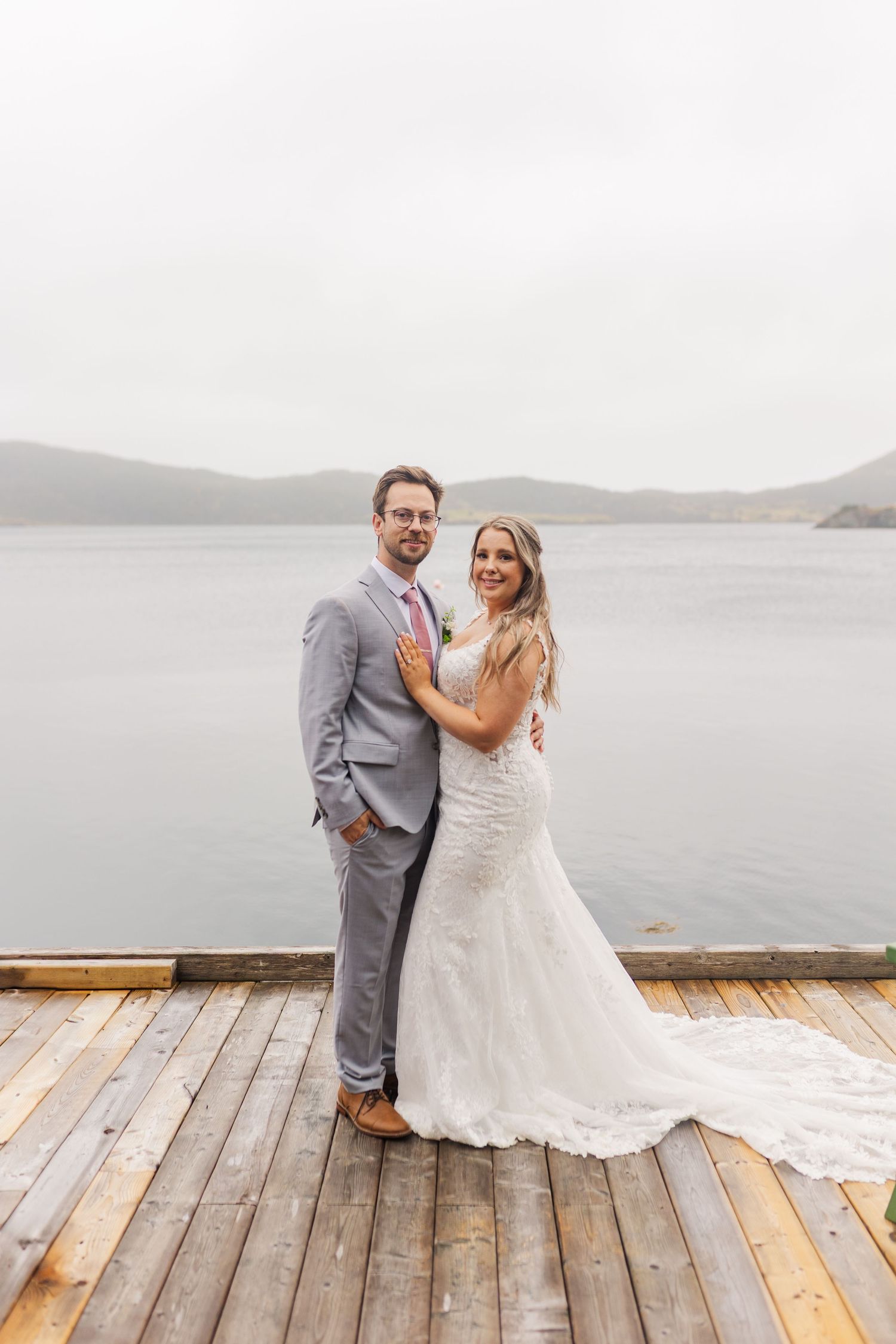 A couple stands together on a wooden dock overlooking a serene lake with mountains in the background.