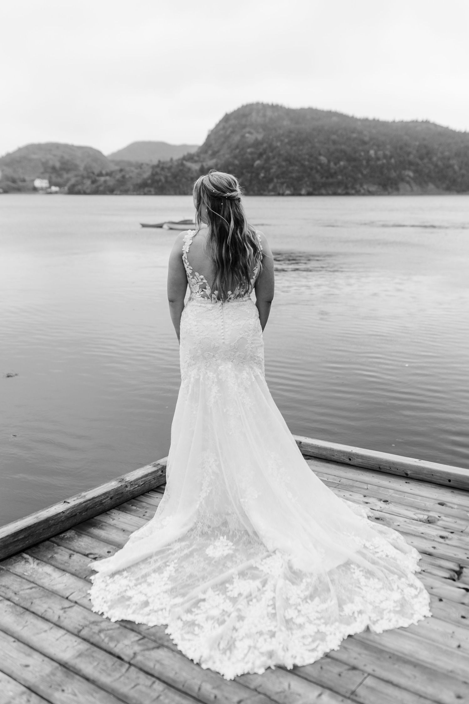 Detailed lace wedding dress train spreads across wooden dock with water and mountains in background.