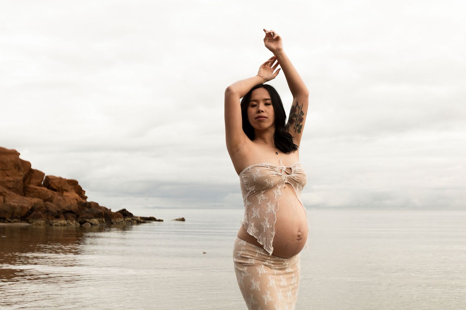 A person in a flowing beige lace dress stands gracefully along a rocky beach shoreline at sunset.