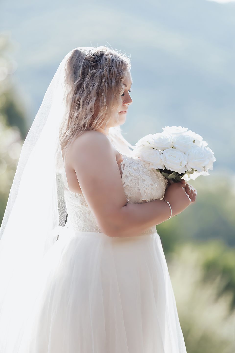 Bride in white lace gown with flowing veil holds bouquet against mountain backdrop.