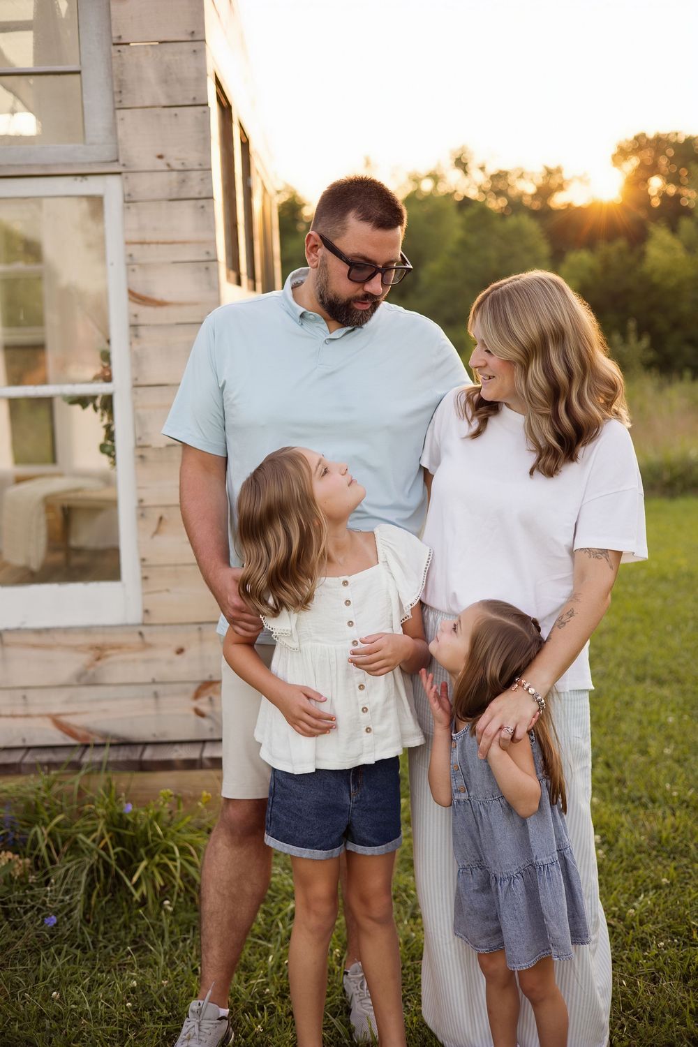 A family of four poses outdoors at golden hour near a rustic building, parents embracing two young daughters.