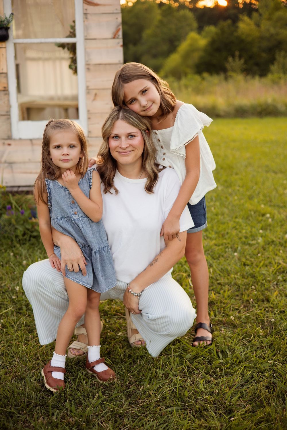 A mother kneels on green grass with her two daughters, posing together near a rustic white structure at sunset.