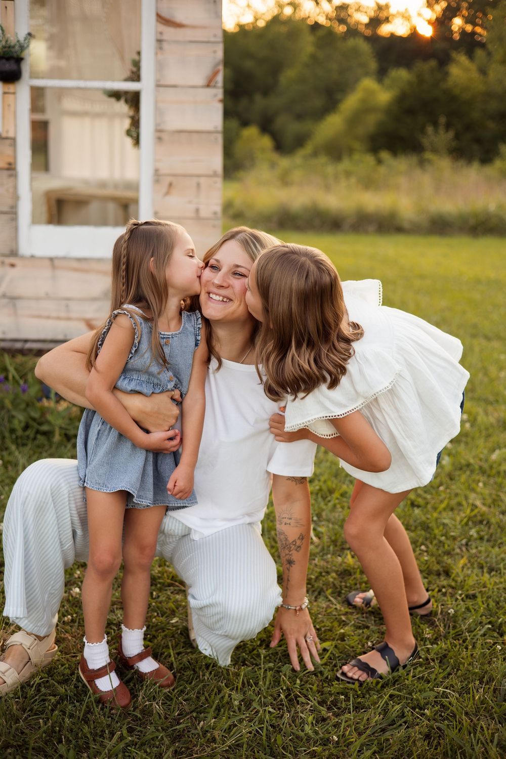 A smiling mother kneels outdoors as two young daughters kiss her cheeks in a sunny green field near a rustic white building.