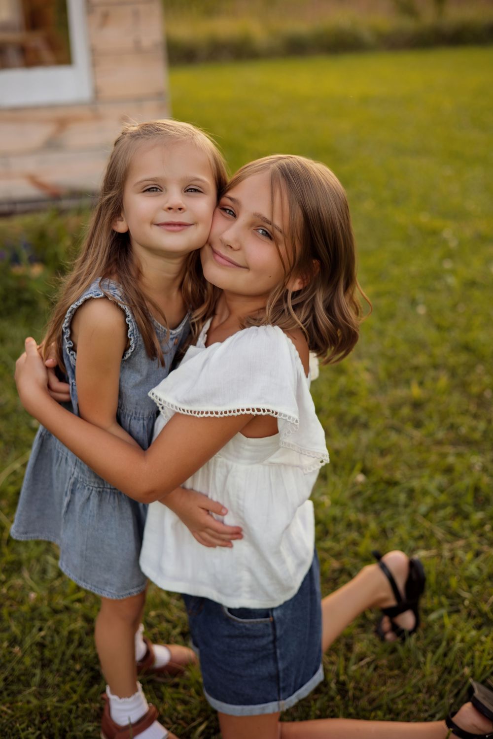 Two young sisters hugging outdoors on green grass, wearing blue and white summer dresses, smiling warmly.