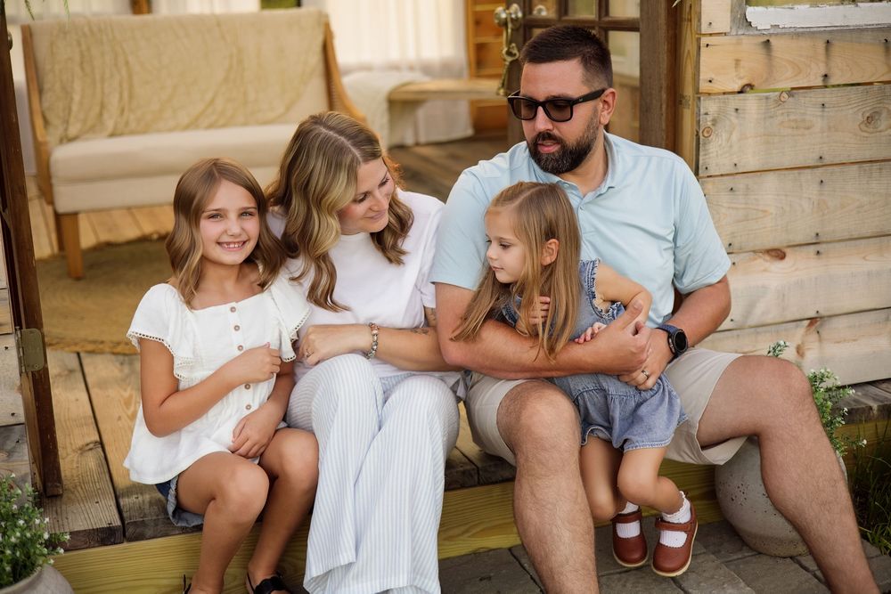 Happy family of four sitting on wooden porch steps, parents with two blonde daughters in casual summer outfits.