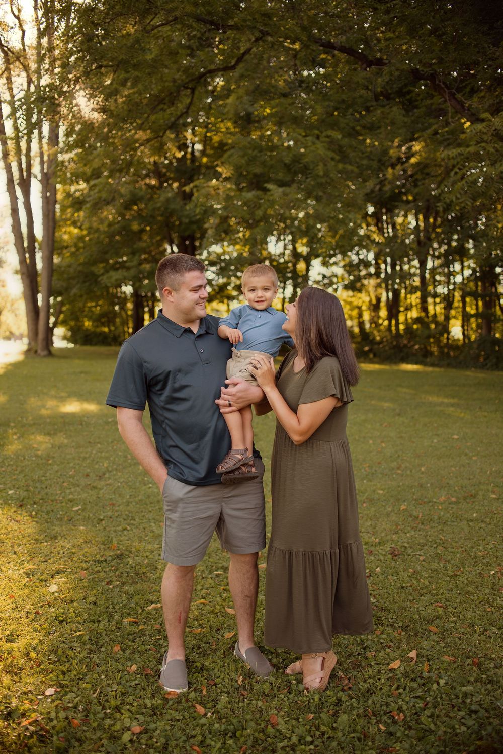 A family stands together in a sun-dappled park setting during golden hour, surrounded by tall trees and green grass.