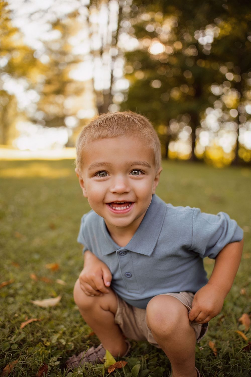 A young child in a gray polo shirt crouches and plays outdoors during golden hour lighting.