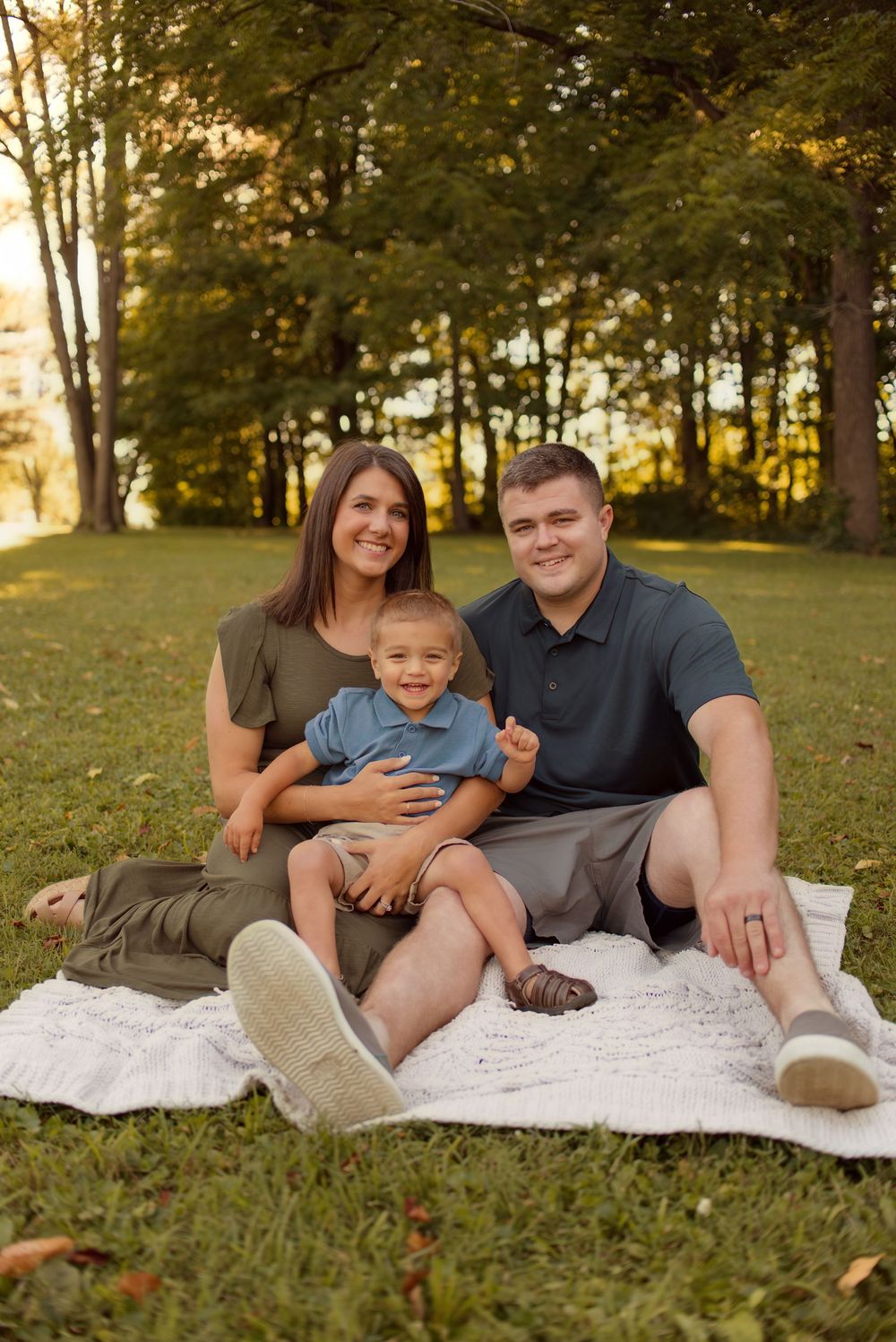 A family sits together on a white blanket in a grassy park during golden hour, sharing warm smiles and embraces.