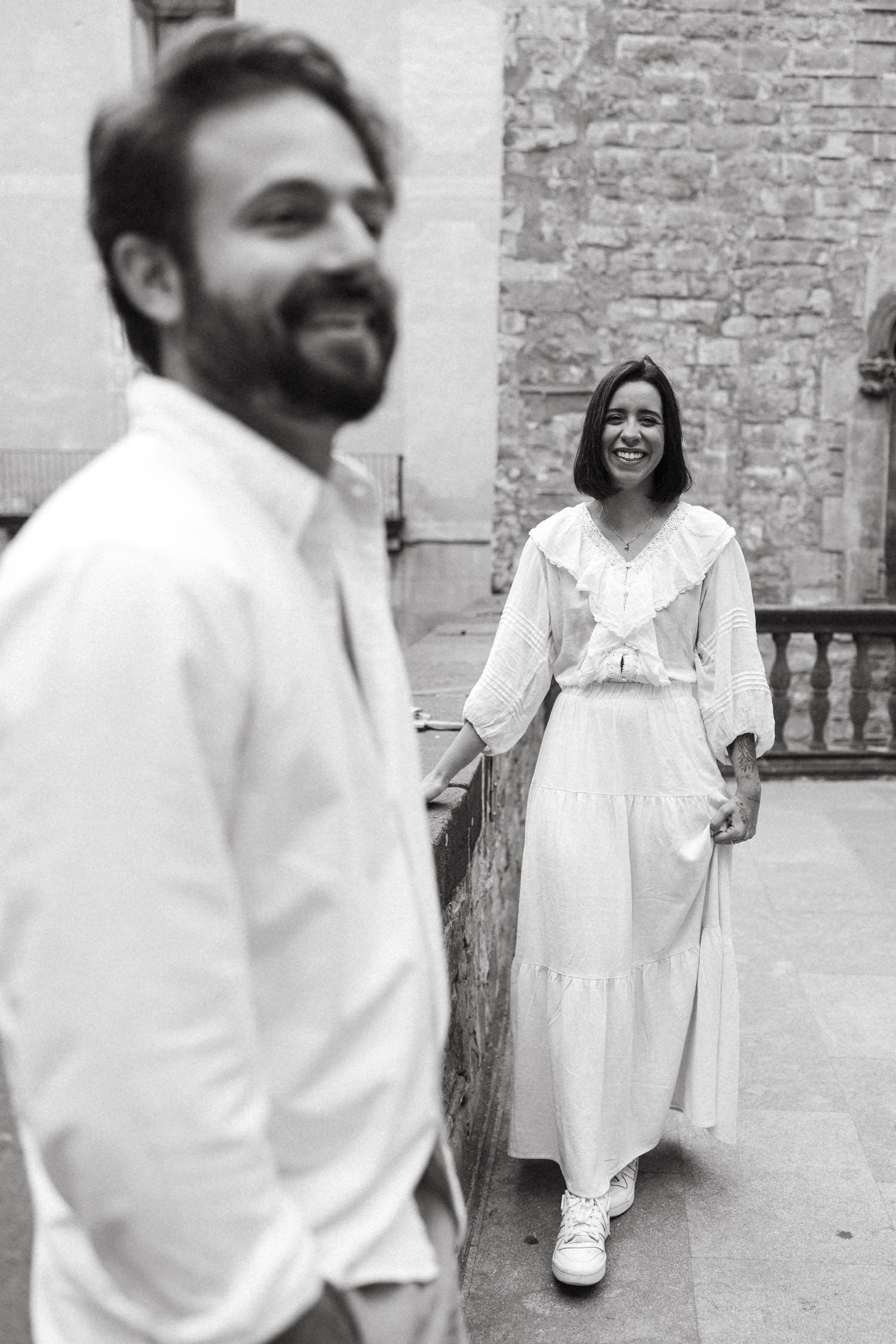 A black and white portrait of a couple in white outfits posing against stone architecture.
