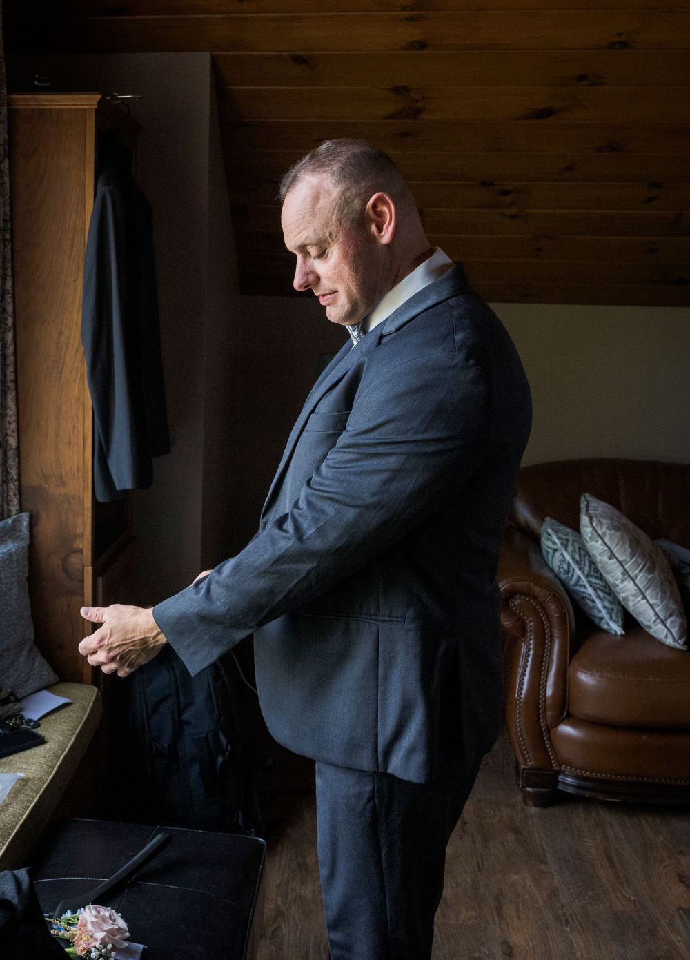 A person in a dark suit checks their phone while standing in a rustic wooden room with leather furniture.