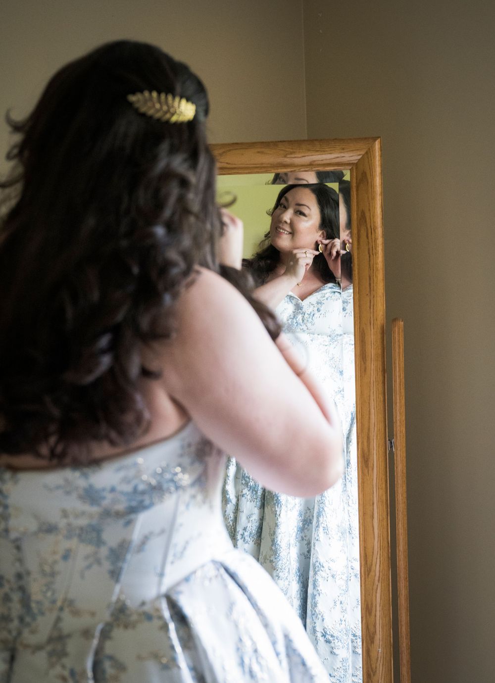 Person with dark curly hair wearing a floral patterned top stands in front of a mirror and window in a sunlit room.