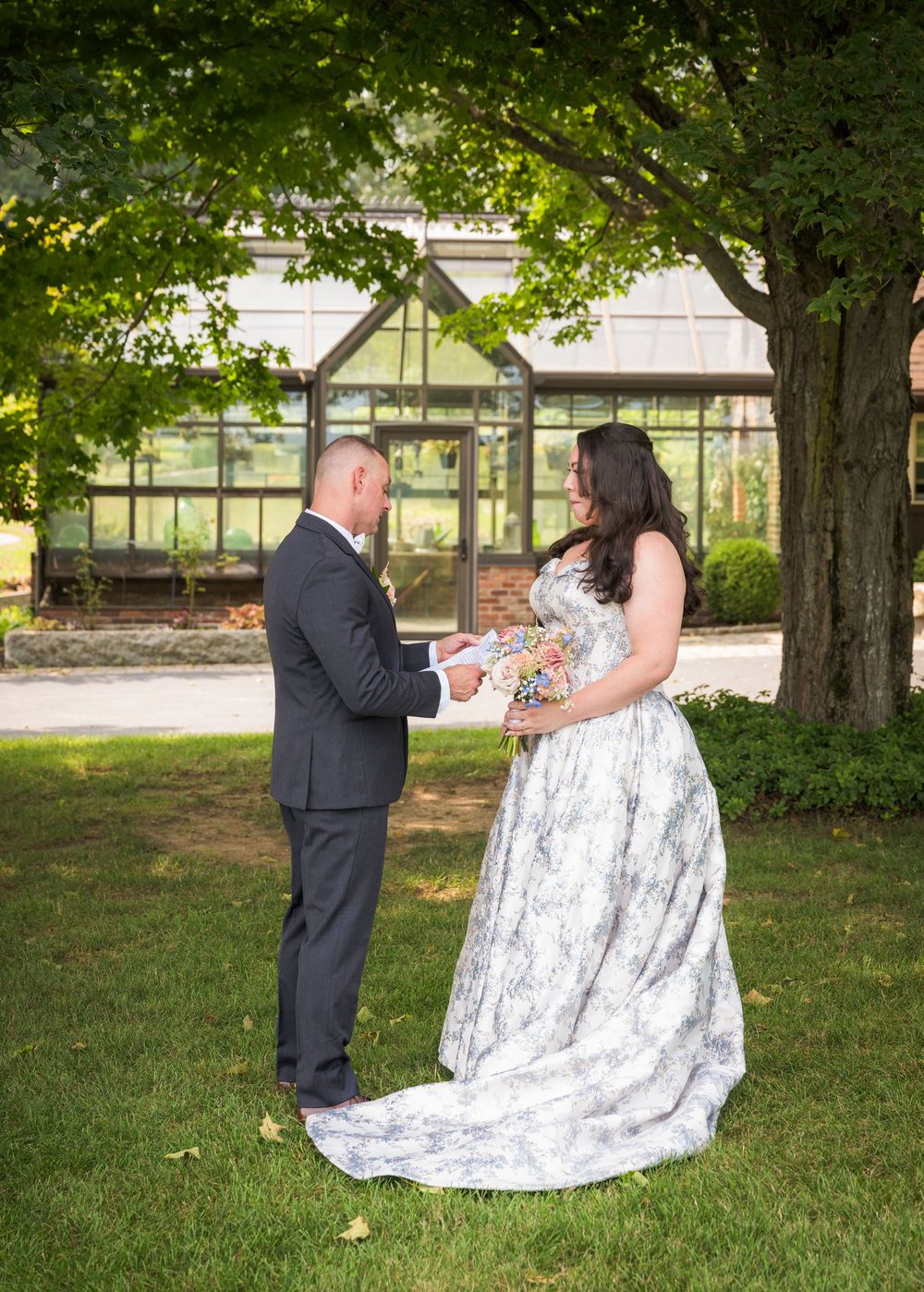 A couple in formal attire shares a romantic moment outdoors beneath leafy green trees on a sunny day.